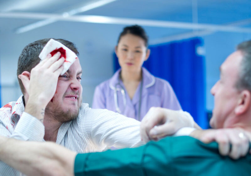 A aggressive patient in an emergency department becomes physically aggressive towards a paramedic, while a nearby nurse appears frozen in fear, illustrating the risks of occupational violence and the need for de-escalation and team safety strategies.